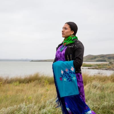 En amerikansk ursprungsbefolkningskvinna står på en gräsbevuxen slätt med ett vattendrag i bakgrunden. Hon är klädd i traditionella kläder och man ser henne i profil titta mot fjärran.