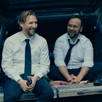 Kaki miesta valkoisissa paidoissa, mustissa housuissa ja kravateissa istuu ruumisauton avoimessa takaosassa. Miehet nauravat. Kuvassa ovat näyttelijät Pekka Strang ja Jari Virman.