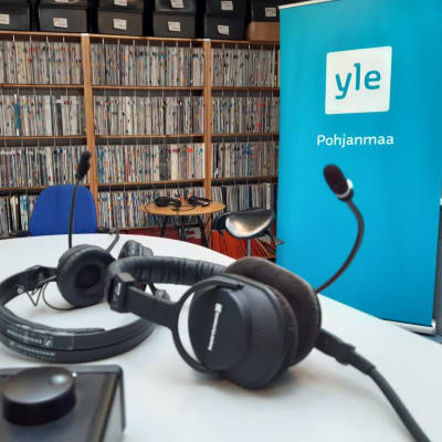 Yle Pohjanmaan studiossa pöydällä kuuloke-mikrofoni ja taustalla levyhylly sekä Yle Pohjanmaa -ständi.