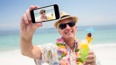 äldre man med solhatt tar en selfie på stranden, har en drink i handen.