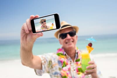 äldre man med solhatt tar en selfie på stranden, har en drink i handen.
