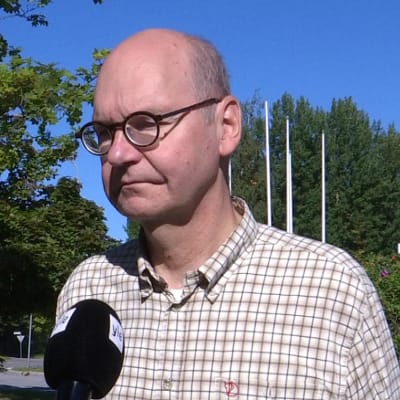 En flintskallig man med glasögon och rutig skjorta står i solen med en mikrofon framför sig.
