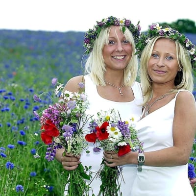 Två unga kvinnor står på en blommande äng med midsommarkransar i håret.