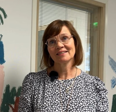 Silmälasipäinen nainen hymyilee kameralle naivistisen seinämaalauksen edessä.