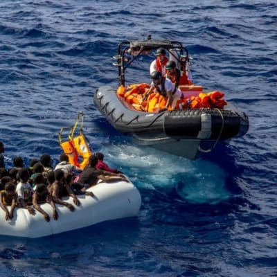 En gummibåt med flyktingar nås av en annan gummibåt med räddningsmanskap som kastar flytvästar till flyktingarna.