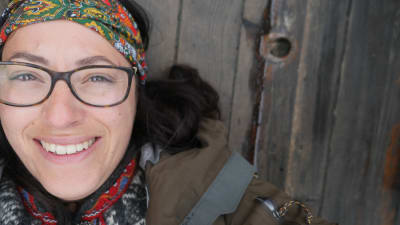 En kvinna med glasögon och en scarf knuten runt huvudet. Ligger på ett träunderlag och tittar in i kameran. 