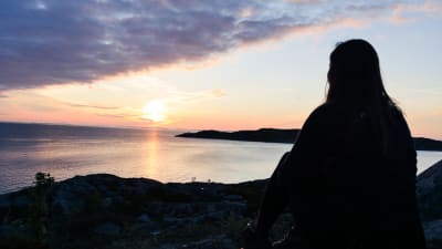 Siluetten av en kvinna som sitter på en klippa och tittar på solnedgången.