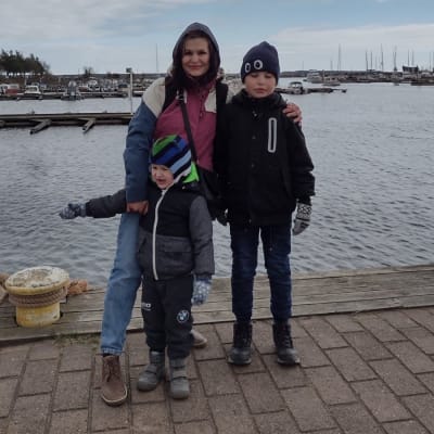En kvinna med sina två barn vid en kaj med hamn i bakgrunden.