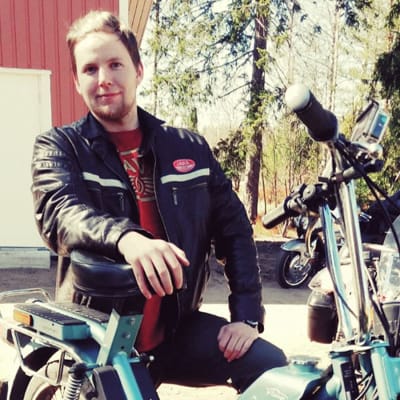 En man med blont hår och läderjacka står bredvid en gammal moped. 