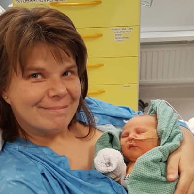 Mamma med nyfött spädbarn i armarna. De ligger på en sjukhusbädd. Mamman ler och bebisen sover.