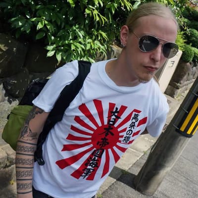 En man med långt blont hår. Han bär solglasögon och en t-shirt med med japansk text på.