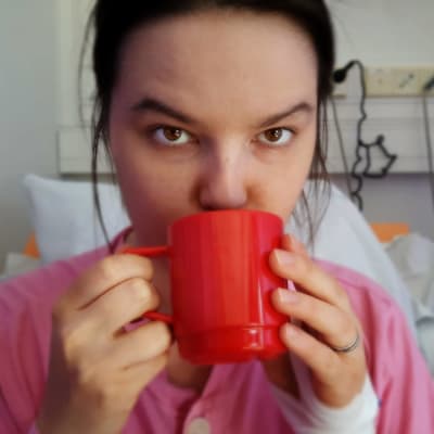 Pernilla Bergman sitter i sin sjukhussäng, ser in i kameran och dricker ur en röd mugg