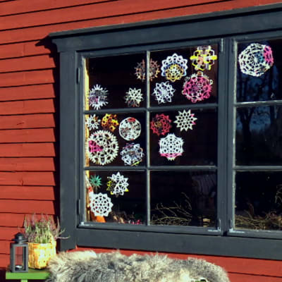 Ett fönster med svart foder, karm och spröjs. Fönstret är dekorerat med nästan tjugo papperssnöflingor i olika mönster och färger. Framför fönstret står en bänk med ett fårskinn, en svart stallykta och en spånkorg med ljung. Bilden är tagen från utsidan.