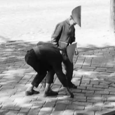 Kaksi pahvihattuista poikaa yrittää irrottaa katuun liimattua seteliä ohjelmassa Rahaa.