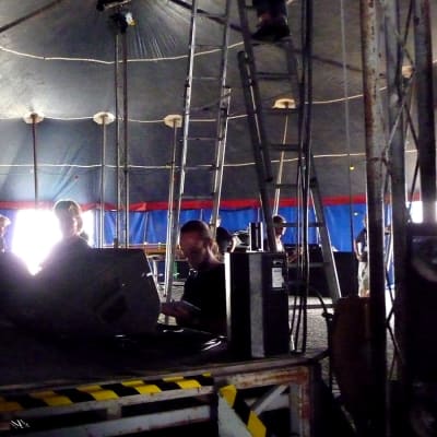 Esiintymislava teltan sisällä, miehiä kasaamassa kaiuttimia ja valoja lavalle. 