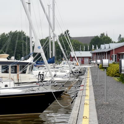 Flera båtar står förtöjda vid en brygga i en hamn. I bakgrunden syns ett rött hus.