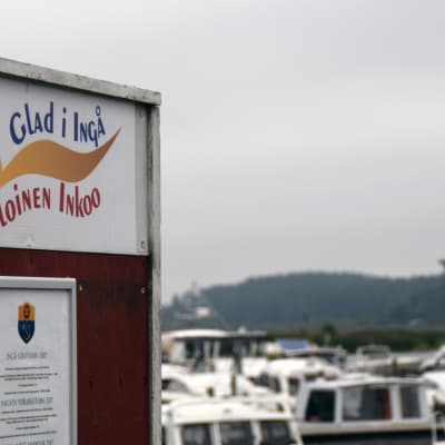 En skylt där det står Glad i Ingå på svenska och finska. I bakgrunden syns båtar i en småbåtshamn,