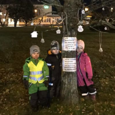 Barn runt ett träd som har reflexer hängandes från grenarna. Trädet är på en gräsmatta och barnen är klädda i vinterkläder. Det är mörkt ute.