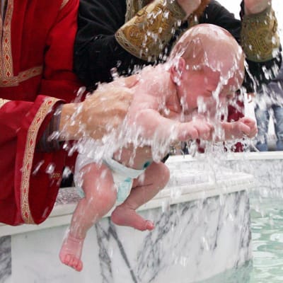 Flera män som är klädda i prästkåpor. Två spädbarn doppas i en stor bassäng under ett dop i Georgien.  