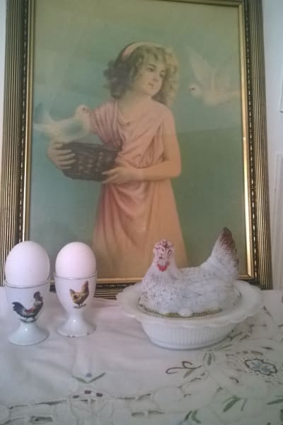 En tavla i bakgrunden föreställande en flicka med två vita duvor. I förgrunden två äggkoppar och en vit porslinshöna.