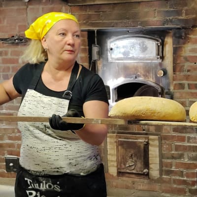 Nainen ottaa leipälapiolla paistuineita leipiä ulos leivinuunista.