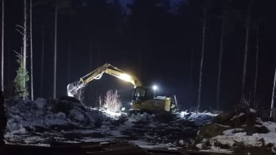 En gul grävmaskin jobbar i mörkret. i bakgrunden syns träd.