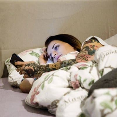 En ung kvinna ligger i sängen och tittar på sin mobiltelefon.