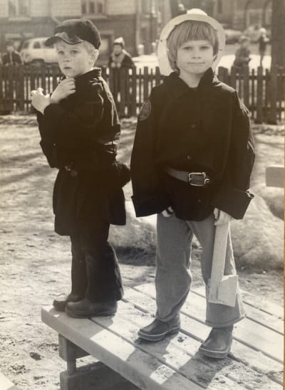 Nokikolariksi ja palomieheksi pukeutuneet Aapo Rista ja Tuomas Heikkilä seisovat leikkipuiston pöydällä, taustalla aita ja sen takana isompia lapsia.