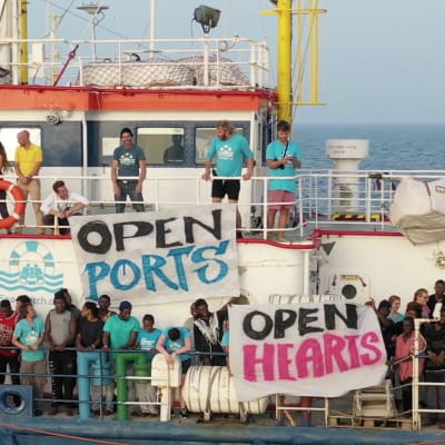 En bild på båten Sea Watch med flyktingar ute på däcket med skyltar där det står "open ports" och "open hearts".