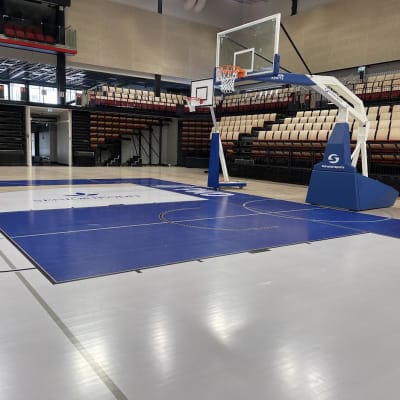 Kuva Joensuun Mehtimäen uudesta palloiluhallista, Motonet-areenasta. Kuvassa oikealla on iso ja pienempi sinivalkoinen koripallokori. Myös lattia on sinivalkoinen. Taustalla katsomoita. 
