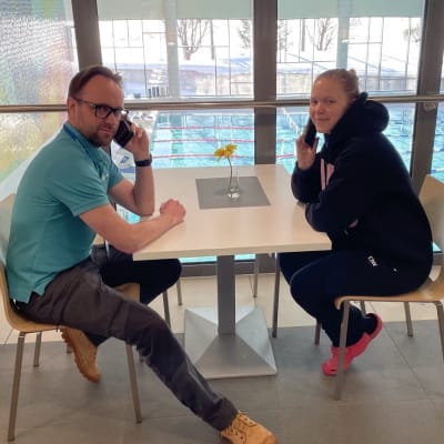 Pöydän ääressä istuvat mies ja nainen puhuvat puhelimessa. Taustalla näkyy uimahallin uima-allas.
