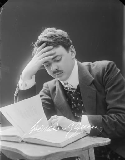 Mustavalkoisessa kuvassa viiksekäs mies lukee kirjaa käsi otsallaan.