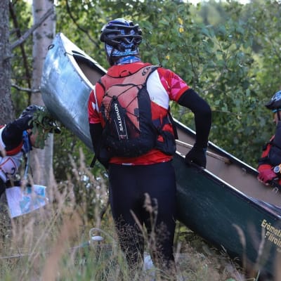 Kolme seikkailukisan osallistujaa kantaa kanoottia kaislikossa.