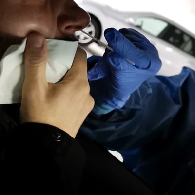 Sjukskötare i skyddsmundering sträcker in en provtagningssticka genom bilfönstret. En man med nerdraget munskydd blir coronatestad.