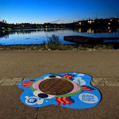 Kaivonkansi ja sen ympärillä oleva värikäs tarra Töölönlahdella ilta-aikaan. Tarrassa on kuva suu auki olevasta kalasta.