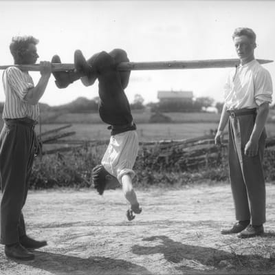 En man leker "flå katt", där han hänger upp och ner på en stång och ska ta sig framåt med att "hoppa" med benen. Bilden är tagen 1930 i Jomala, Björsby, Åland.