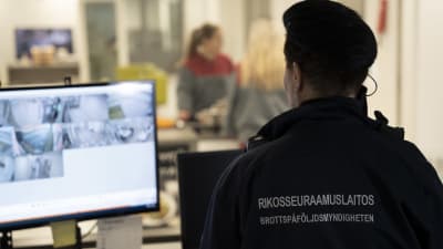 Vanginvartija istuu selin kameraan vieressään valvontamonitori ja edessään kaksi epätarkasti näkyvää naisvankia.