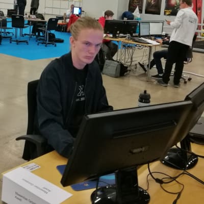 Ung svartklädd man sitter framför två datorskärmar i en stor sal där det också finns andra människor.