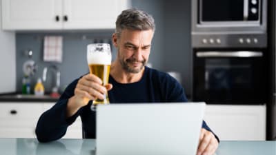 Man sitter i ett kök och skålar med en öl mot en datorskärm. 