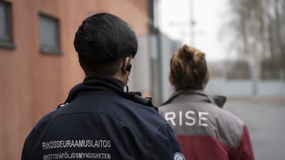 Kaksi naista kävelevät peräkkäin betonirakennuksen pihalla. Takaapäin otetussa kuvassa näkyy, että edellä kävelevän vangin vaatteen selässä lukee RISE ja takimmaisena tulevan vartijan Rikosseuraamuslaitos.