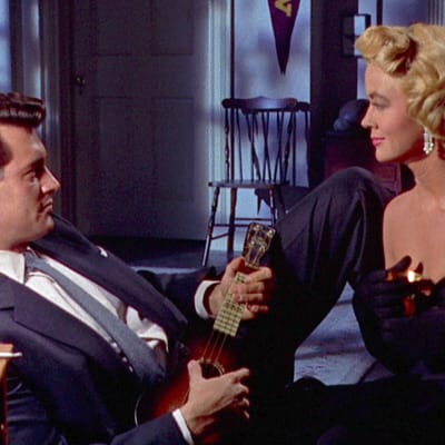 Mies (näyttelijä Rock Hudson) puvussa ja solmiossa nojailee sohvalla ukulele käsissään ja katsoo hymyillen olkaimettomassa mekossa hänen edessään istuvaa vaaleaa naista (näyttelijä Dorothy Malone), kuva elokuvasta Tuuleen kirjoitettu.