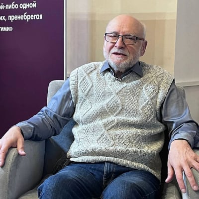 Harmaapartainen silmälasipäinen mies,  Vjatšeslav Bahmin, istuu nojatuolissa ja puhuu hieman hymyillen. Taustalla huoneessa näkyy juliste.