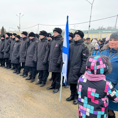 Joukko sotilaita seisoo rivissä aukiolla väkijoukon keskellä. Yhdellä sotilaalla on valko-sininen lippu kädessään.