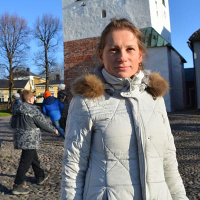 En kvinna i vit jacka står framför klocktornet vid Borgå domkyrka. Bakom henne syns två lekande barn och en kvinna i svart jacka som går förbi.