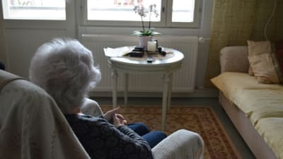 Äldre kvinna ser ut genom fönster.