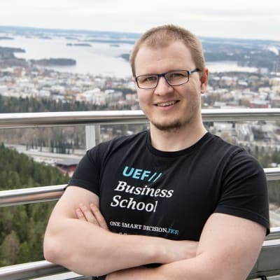 Ung man i svart t-shirt och glasögon står på utsiktsplats, i bakgrunden en stad vid en sjö på långt avstånd.
