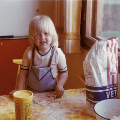 Iiu Susiraja lapsena pöydän ääressä leipomassa. Pöydällä on iso vehnäjauhopussi ja keltainen muovipurkki.