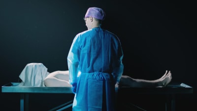 Leikkauspöydällä makaa ihminen, jonka kasvot on peitetty liinalla, pöydän edessä seisoo toinen ihminen suojavarusteissa.