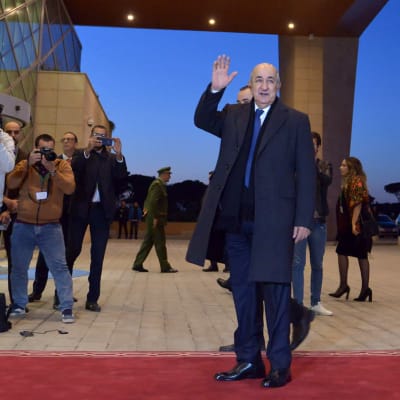 Algeriets president Tebboune vinkar till kameran. Till vänster flera fotografer.
