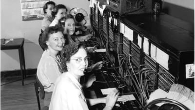 en bild av fem kvinnor som arbetade som telefonoperatörer på femtiotalet. framför sig har de paneler med kablar för att koppla samtalen med.
